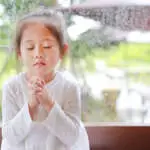 Adorable little Asian girl praying at glass windows on the raining day. Spirituality and religion.