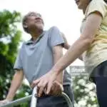 Asian happy Senior retired grandfather enjoy outdoor at green park. Attractive girl daughter help elderly handicaps father walk using walker or cane for hearth care activity. Medical Insurance concept