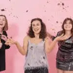 Beautiful cheerful festively dressed girls on pink background with fireworks, candles and confetti, new year party concept.