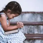 Cute asian little girl closed her eyes and  folded her hand in prayer on a Holy Bible for faith concept in vintage color tone