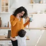 Cute little boy embraces his mom  at kitchen while she is talking by phone holding cup, smiling, looking at son. Cheerful hispanic young woman in orange  blouse happy at home with beloved little boy.