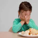 Cute little girl crying and refusing to eat vegetable salad at table on grey background