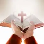 Hands of Woman Raise up a Bible for Praying with Blurred Shape of Cross and Heart