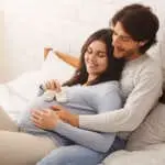 Happy young couple expecting baby. Pregnant millennial woman and her husband lying on bed with little shoes near belly, enjoying future parenthood