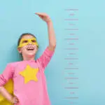 Little child plays superhero. Kid measures the growth on the background of bright blue wall. Girl power concept. Yellow, pink and  turquoise colors.