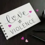 LOVE ≠ VIOLENCE black lettering in notebook with pink paper hearts on desk