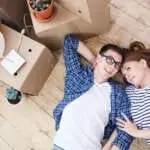 Lovely young couple lying on wooden floor of their new apartment and sharing  ideas concerning interior design of living room, pile of moving boxes standing next to them