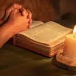 Man's hands in prayer posture on a bible with candle lighting