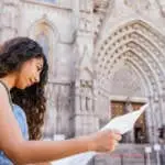 Side view of young Hispanic female traveler with long curly hair examining paper map while standing on street outside cathedral during visit in historic city
