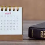 Spiral calendar for April month 2023 with date numbers and closed holy bible book on wooden table. A close-up.