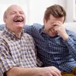 Two men father and son are sitting on the sofa in the living room at home and remember the funny events from the past. They laugh at the joke. Good family relationships