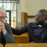 Young black man in shirt with clerical collar keeping hand on shoulder of senior male parishioner while comforting him and praying