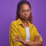 Young offended African American woman pouts lips and turns away to show displeasure or resentment stands with arms crossed in front of chest posing on plain purple background. Ethnic portrait