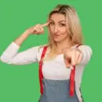 You're idiot with insane crazy plan. Portrait of displeased adult woman in denim overalls showing stupid gesture and pointing to camera, accusing of dumb idea. studio shot isolated on green background