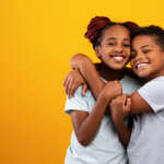 Emotional african american teenagers brother and sister twins or siblings in similar white t-shirts embracing and smiling at camera over yellow studio background, panorama with copy space