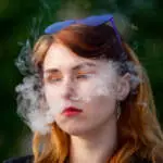 Vape teenager. Young pretty white caucasian girl with glasses on her head an electronic cigarette on the street in the summer. Deadly bad habit.