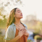 Young latin woman with hand on chest breathing in fresh air in a beautiful garden during sunset. Healthy mexican girl enjoying nature while meditating during morning exercise routine with closed eyes. Mindfulness woman enjoying morning ritual and relaxing technique.