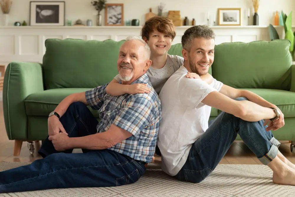 Joyful multigenerational male family sitting on warm floor in living room. Happy schoolboy enjoying weekend with smiling grandpa and young handsome dad at home. Father's day and parenthood concept