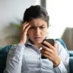 Confused millennial Indian girl sit at home look at cellphone having operational problems, frustrated young woman feel stressed with slow Internet connection, virus attack or spam on smartphone