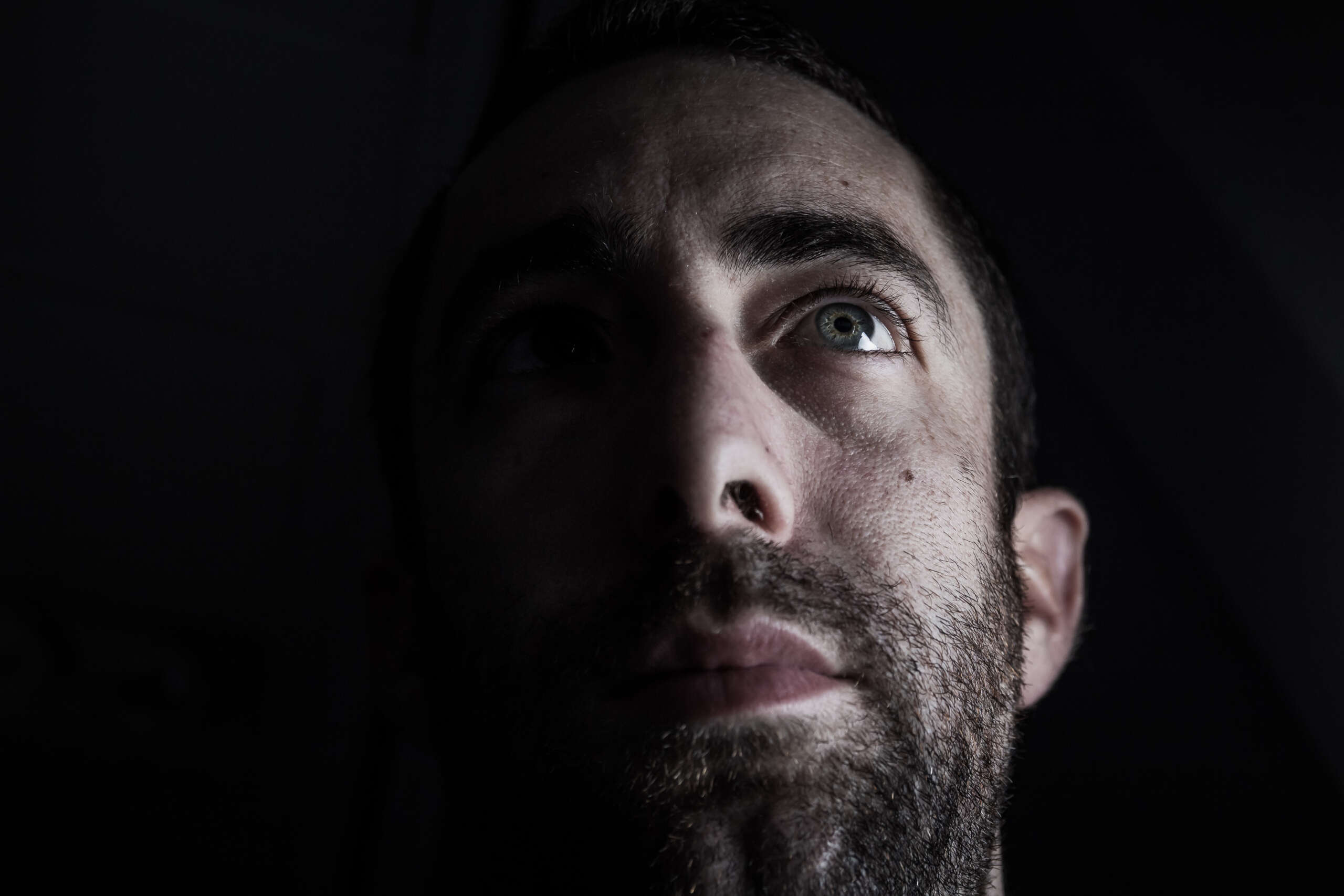 Dramatic portrait of a young man half expressionless face in deep shadow looking up isolated on black background.