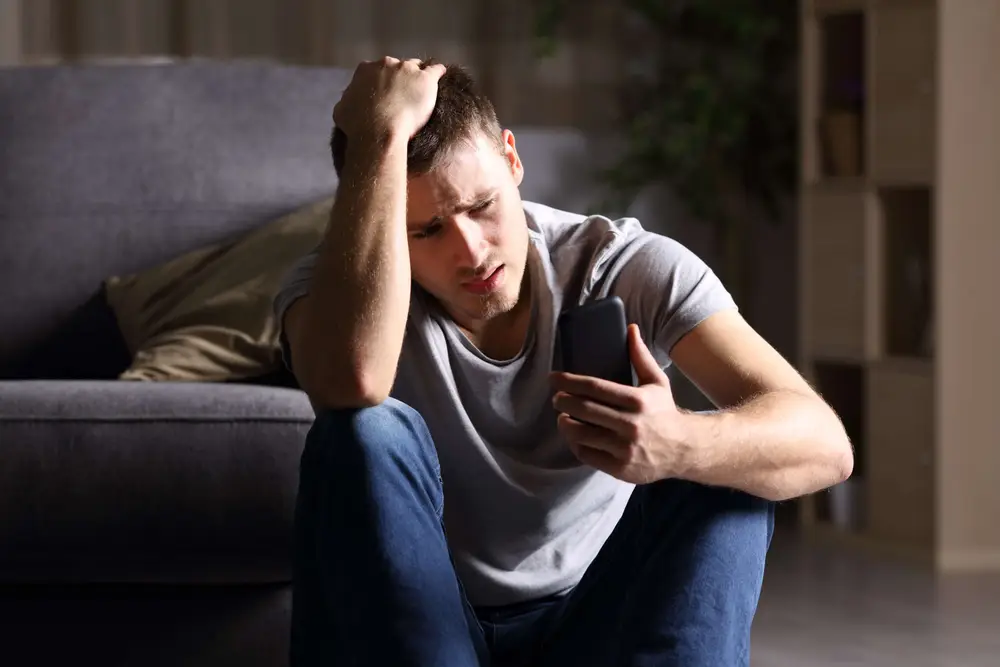 Single sad man checking mobile phone sitting on the floor in the living room at home with a dark background