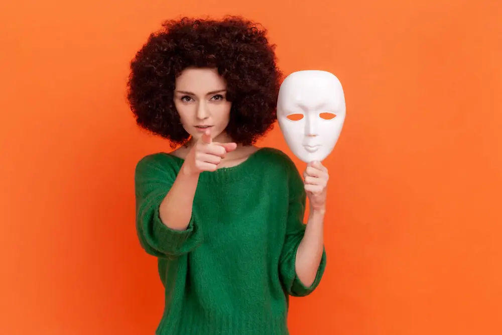 Strict bossy woman with Afro hairstyle in green casual style sweater holding white mask in hand, pointing finger to camera with serious expression. Indoor studio shot isolated on orange background.